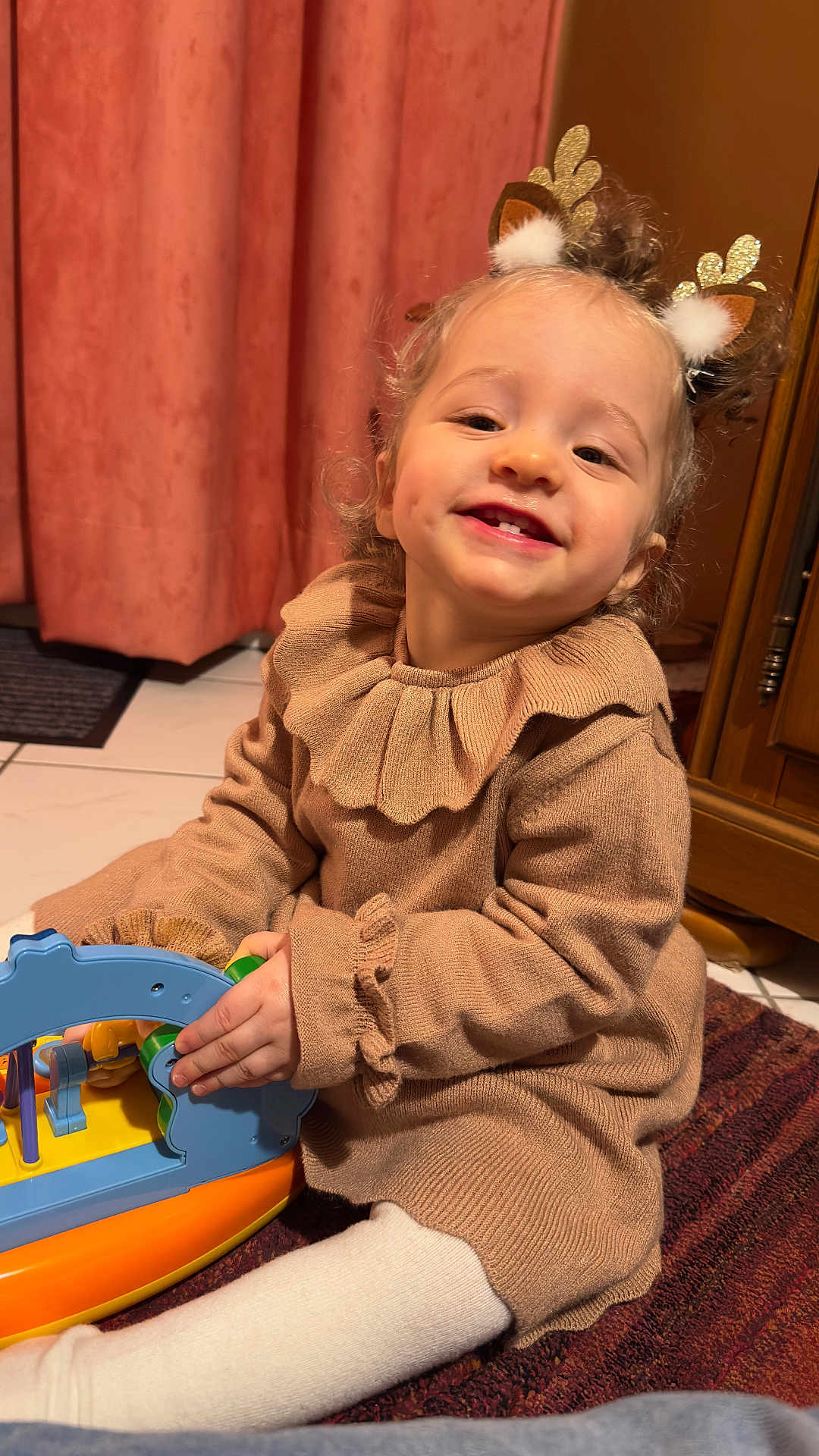Chiara a rejoint le concours — aidez-le/la à gagner de superbes lots ! child, toddler, smiling_child, toy, headband, antler_headband, sweater, beige_clothing, curtain, indoor, playtime, hands, dimple, white_tights, carpet, tile_floor, wooden_cabinet, close_up, warm_lighting, happy_face