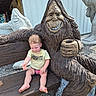 barefoot, bench, bigfoot, building, casual_clothing, child, curious, daylight, gravel, holding, mythical_creature, outdoor, person, rustic, sculpture, smiling, statue, toddler, wood, young_child