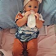 Haylee is registered to the contest to win money with this photo: baby, bed, blanket, bottle, bow, child, clothing, drink, face, feeding, footwear, headband, infant, lying_down, orange_pants, pillow, portrait, striped, toddler, wearing