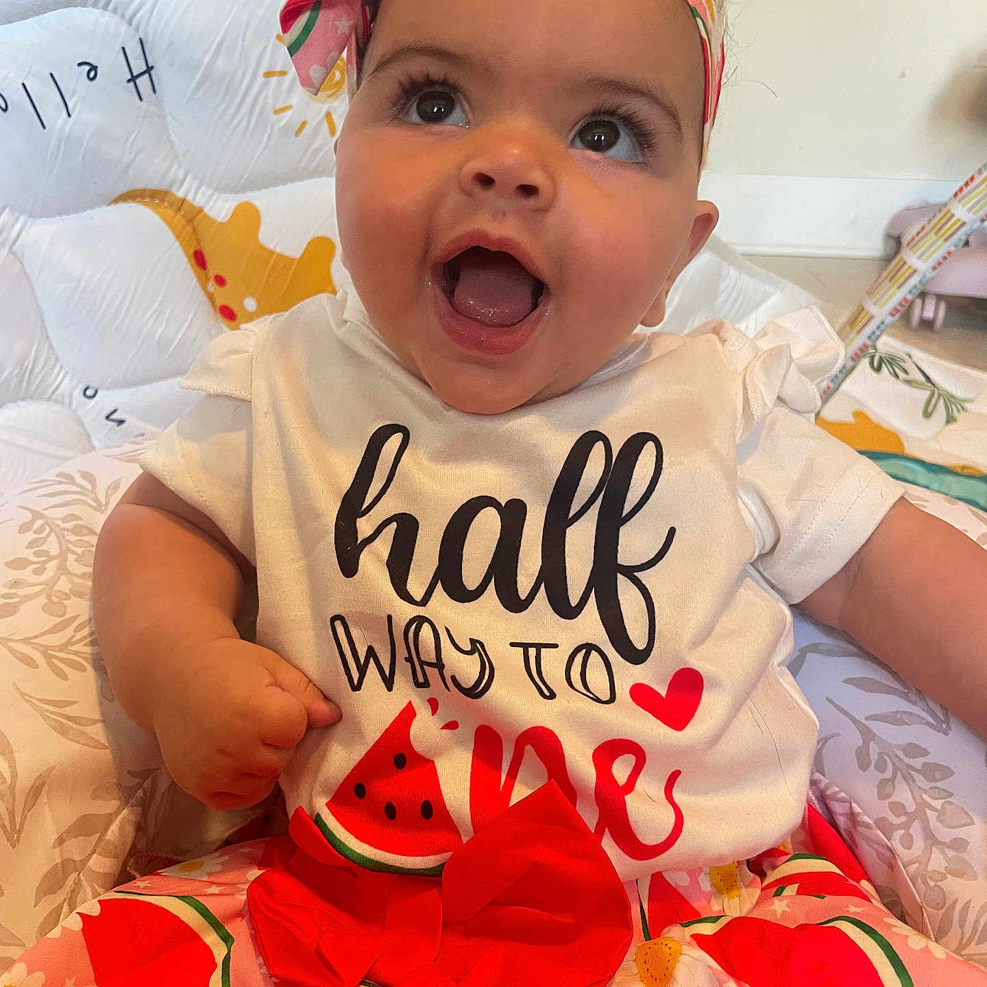 Wynter is registered to the contest to win money with this photo: baby, child, smiling, happy, headband, watermelon, dress, flower, patterned_clothing, cushion, indoor, portrait, cute, infant, sitting, bright_colors, face, expression, toddler, playful