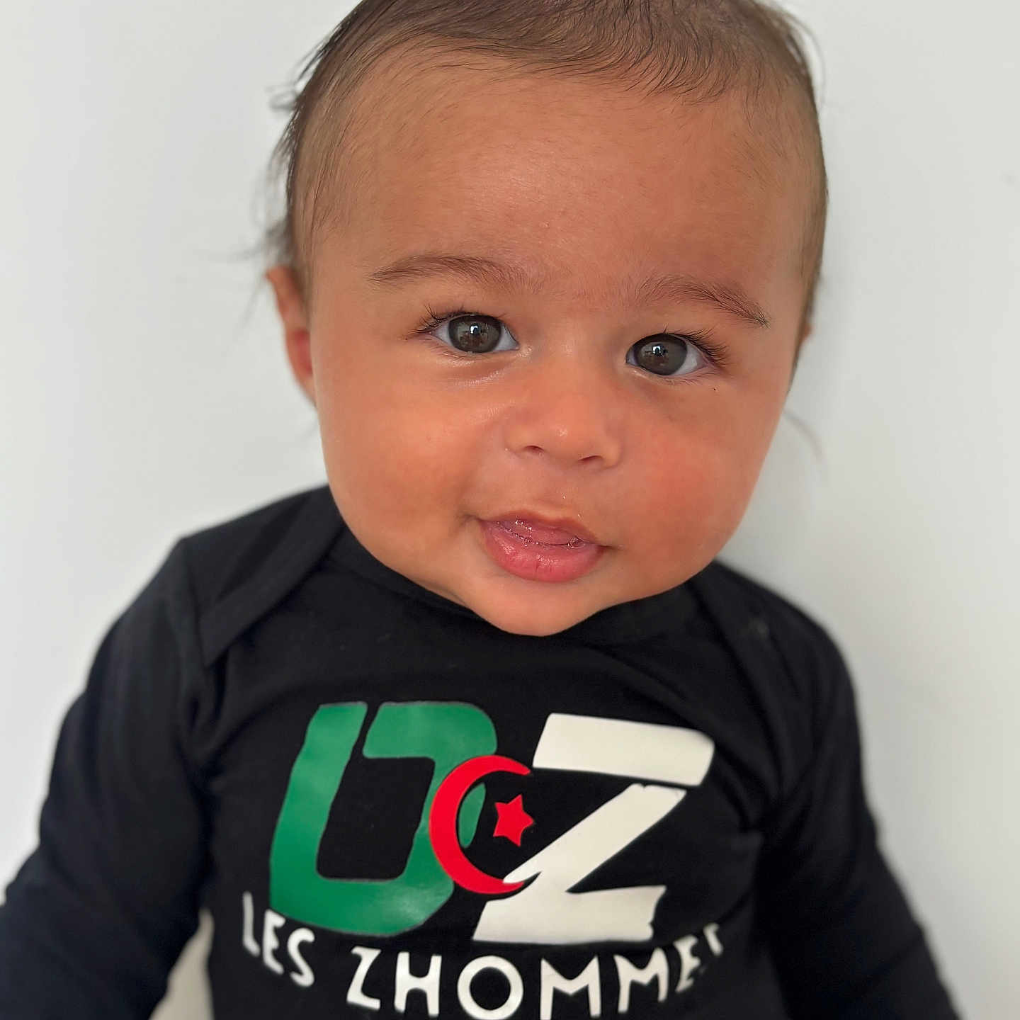 Nassim Djellouah participe au concours pour gagner de l'argent avec cette photo : baby, black_shirt, casual_clothing, child, colorful_text, curious, cute, expression, eyes, face, head, indoors, infant, long_sleeve, portrait, sitting, skin, smile, white_background, young_child