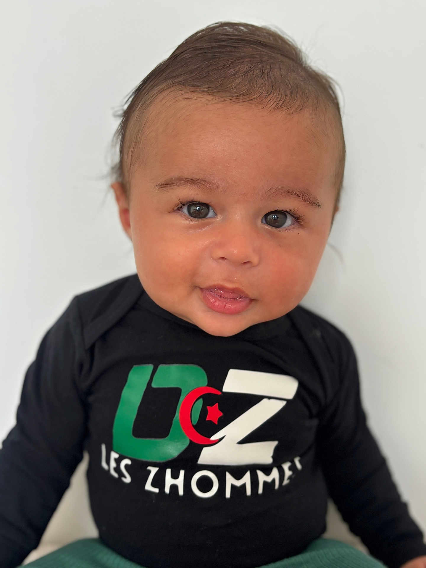 Nassim Djellouah participe au concours pour gagner de l'argent avec cette photo : baby, infant, child, portrait, face, eyes, smile, black_shirt, colorful_text, sitting, white_background, cute, young_child, curious, indoors, long_sleeve, casual_clothing, head, skin, expression