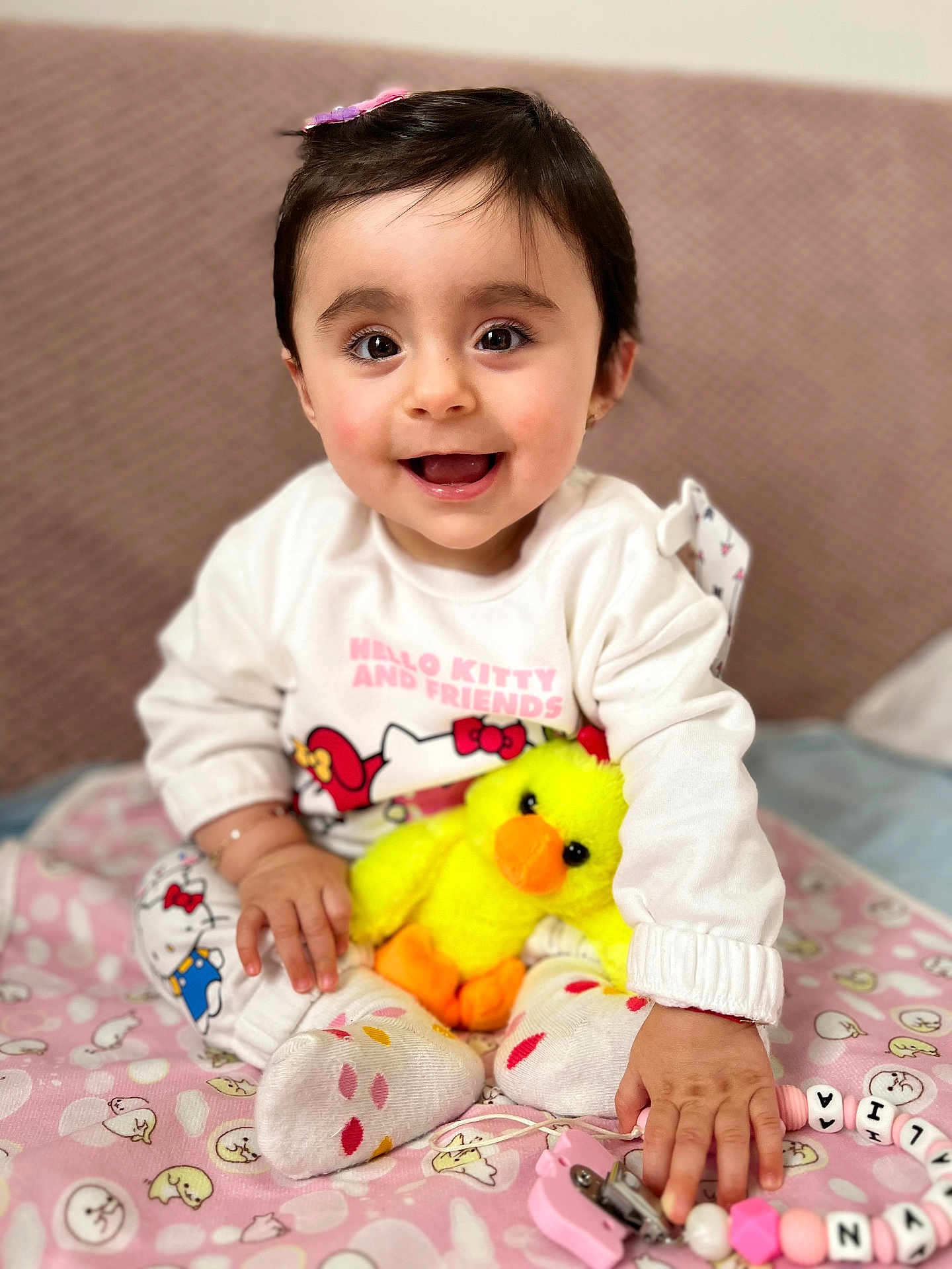 Natalia is registered to the contest to win money with this photo: baby, child, smiling, happy, plush_toy, yellow_duck, hello_kitty, clothing, socks, blanket, indoor, cute, portrait, person, toy, pink, pattern, hand, face, hairclip