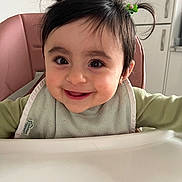 Natalia is registered to the contest to win money with this photo: baby, smiling, high_chair, pigtails, green_hair_clip, bib, indoor, kitchen, child, cute, face, person, happy, young_child, feeding, infant, clothing, seat, portrait, home