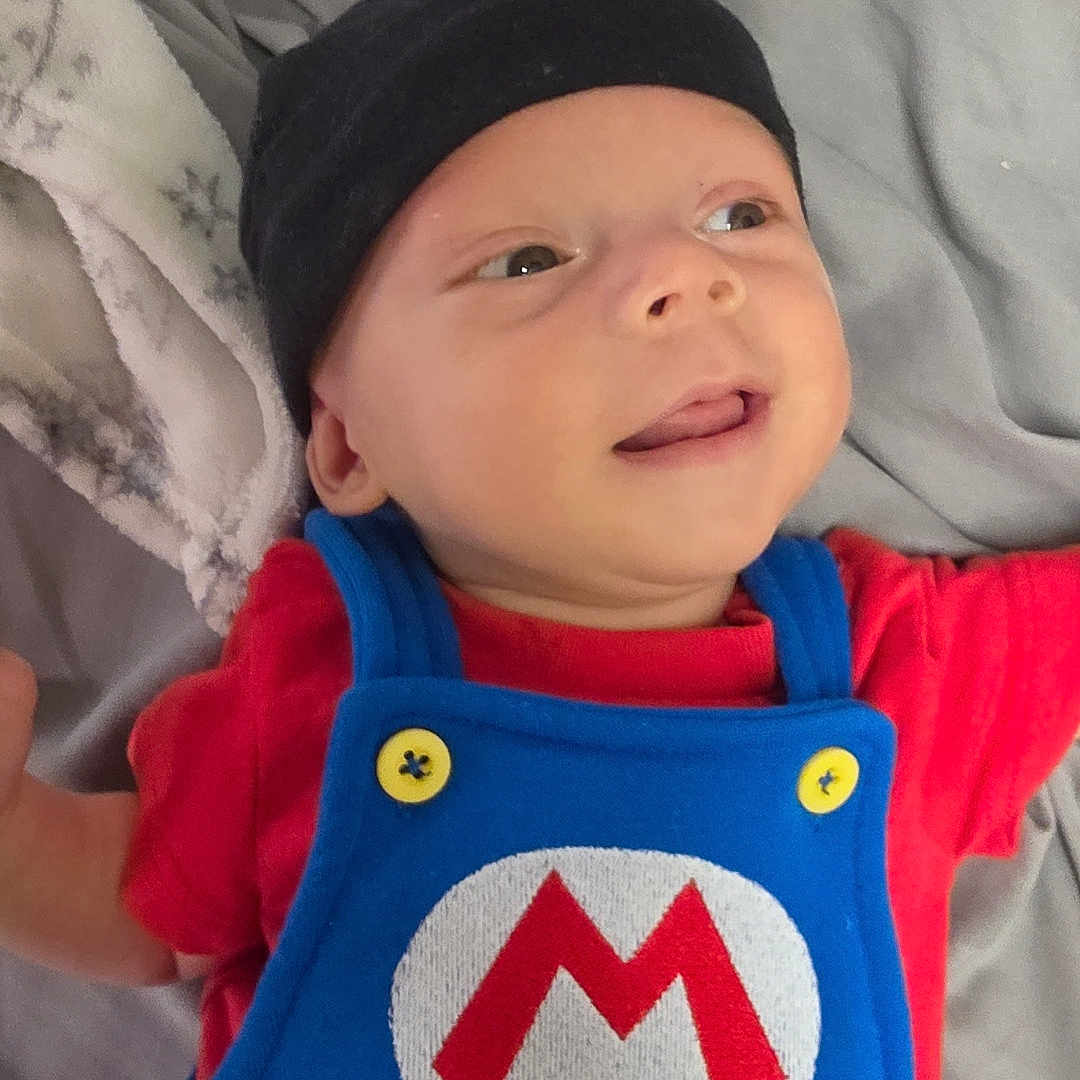 Deidara joined the competition — help win amazing prizes! baby, infant, smiling, blue_overalls, red_shirt, black_hat, hand, tattoos, blanket, bed, cute, person, child, portrait, lying_down, soft_texture, clothing, button, indoor, happy