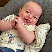 Thomas is registered to the contest to win money with this photo: baby, infant, smiling, tongue_out, mickey_mouse, clothing, quilted_cushion, blanket, blue_shorts, cute, happy, child, face, skin, indoor, portrait, resting, relaxed, person, newborn