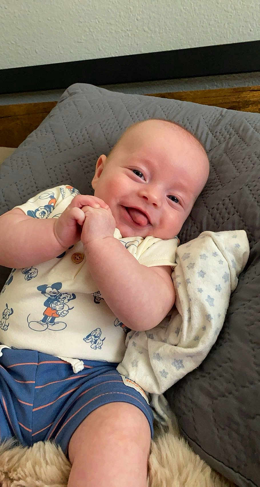 Thomas is registered to the contest to win money with this photo: baby, infant, smiling, tongue_out, mickey_mouse, clothing, quilted_cushion, blanket, blue_shorts, cute, happy, child, face, skin, indoor, portrait, resting, relaxed, person, newborn