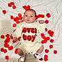 adorable, baby, child, cute, face, hands, heart_pattern, indoor, infant, knitted_outfit, lover_boy, lying_down, portrait, red_petals, red_text, rose_petals, socks, white_clothing, white_sheet, young_baby