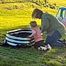 child, adult, grass, garden, soil, outdoor, sunlight, toy_car, kneeling, greenery, planting, playground, backyard, nature, daylight, casual_clothing, activity, family, fun, summer