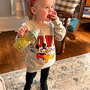 Harlee is registered to the contest to win money with this photo: child, toddler, girl, red_bow, sippy_cup, snack, indoor, living_room, carpet, sweatshirt, mickey_mouse, pluto, black_pants, black_shoes, wood_floor, furniture, couch, cute, portrait, standing