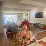 Margaux is registered to the contest to win money with this photo: toddler, child, red_hat, knit_sweater, cherries, candy, kitchen_counter, living_room, toys, couch, wooden_floor, window, curtains, television, carpet, table, air_conditioner, indoor, person, home