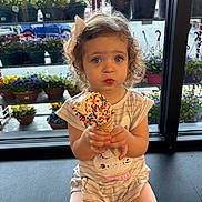 Margaux is registered to the contest to win money with this photo: toddler, child, ice_cream, sprinkles, bow, curly_hair, window, flowers, plant_pots, indoor, sitting, cute, food, dessert, summer, snack, person, expression, clothing, legs