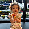 toddler, child, ice_cream, sprinkles, bow, curly_hair, window, flowers, plant_pots, indoor, sitting, cute, food, dessert, summer, snack, person, expression, clothing, legs