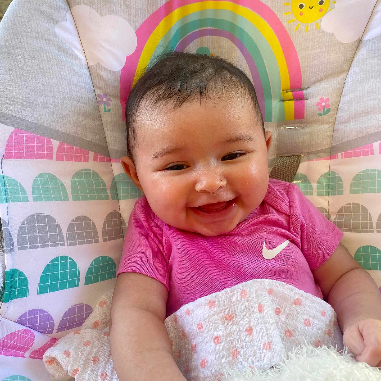 Patsyn is registered to the contest to win money with this photo: baby, blanket, child, close_up, clouds, face, happy, high_chair, indoor, infant, patterned_background, photography, pink_shirt, plush_toy, portrait, rainbow, sitting, smiling, sun, toy