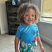 Zakai is registered to the contest to win money with this photo: child, toddler, curly_hair, smiling, blue_tshirt, toy_story, indoor, carpet, dresser, window, blinds, sunlight, happy, casual_clothing, person, portrait, home, young_child, cute, standing