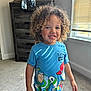 child, toddler, curly_hair, smiling, blue_tshirt, toy_story, indoor, carpet, dresser, window, blinds, sunlight, happy, casual_clothing, person, portrait, home, young_child, cute, standing