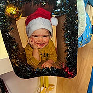 Néo participe au concours pour gagner de l'argent avec cette photo : child, toddler, santa_hat, smiling, holiday, christmas, tinsel, ornament, cardboard, festive, yellow_shirt, indoors, decoration, portrait, happy, cute, face, hand, playful, seasonal
