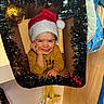 child, toddler, santa_hat, smiling, holiday, christmas, tinsel, ornament, cardboard, festive, yellow_shirt, indoors, decoration, portrait, happy, cute, face, hand, playful, seasonal