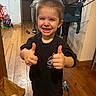 child, smiling, thumbs_up, black_tshirt, indoor, wooden_floor, clothing, laundry_rack, paper_bag, happy, short_hair, person, home, casual, socks, furniture, doorway, backpack, young_child, portrait