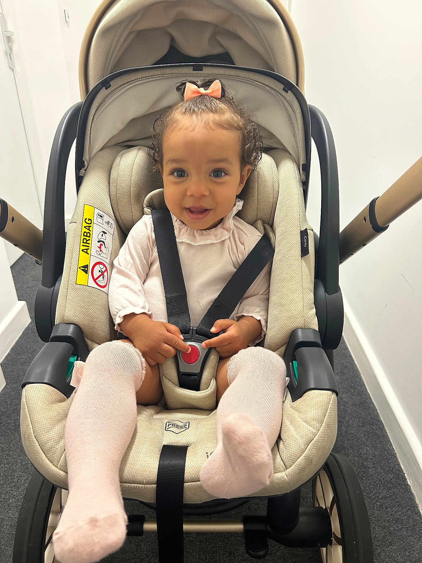 Ariella is registered to the contest to win money with this photo: toddler, child, stroller, car_seat, harness, bow, pink_socks, indoor, carpet, smiling, pointing, baby_clothes, safety, seatbelt, cute, happy, person, infant, baby, transportation