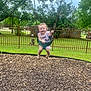 toddler, child, swing, playground, grass, wood_chips, fence, tree, smiling, happy, outdoor, curly_hair, summer_clothing, footwear, nature, recreation, park, daytime, person, fun