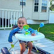 Jeremiah is registered to the contest to win money with this photo: baby, walker, toy, outdoor, deck, grass, house, window, smile, happy, child, blue_clothing, sunlight, foot, person, play, summer, baby_boy, plastic, home
