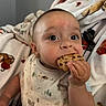 baby, child, cookie, bib, eating, hand, blanket, animal_print, indoor, cute, face, mouth, snack, clothing, person, portrait, infant, messy, curious, happy