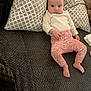 baby, infant, pillow, blanket, patterned_pillow, floral_pants, white_shirt, barefoot, indoors, lying_down, face, person, cute, cozy, soft_texture, curious, resting, home, child, comfort