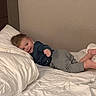 Liam joined the competition — help win amazing prizes! toddler, child, bed, pillow, white_bedspread, blue_shirt, gray_pants, smiling, lying_down, indoor, young_child, person, casual_clothing, resting, playful, home, cozy, room, portrait, cute