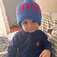 Liam joined the competition — help win amazing prizes! toddler, child, hat, knitted_hat, blue_clothing, bed, bedding, striped, indoors, portrait, cute, face, expression, hand, young_child, cozy, warm_clothing, person, sitting, curious