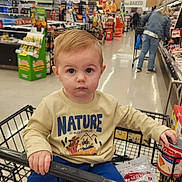 Liam is registered to the contest to win money with this photo: toddler, child, shopping_cart, grocery_store, groceries, clothing, sweatshirt, pants, can, person, indoor, aisle, shopping, supermarket, food, floor, adult, hair, face, expression