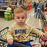 toddler, child, shopping_cart, grocery_store, groceries, clothing, sweatshirt, pants, can, person, indoor, aisle, shopping, supermarket, food, floor, adult, hair, face, expression