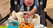 Noah is registered to the contest to win money with this photo: baby, car_seat, blanket, toy, smiling, happy, indoor, child, person, floor, wooden_floor, clothing, seatbelt, colorful, infant, cute, play, expression, young_child, sitting