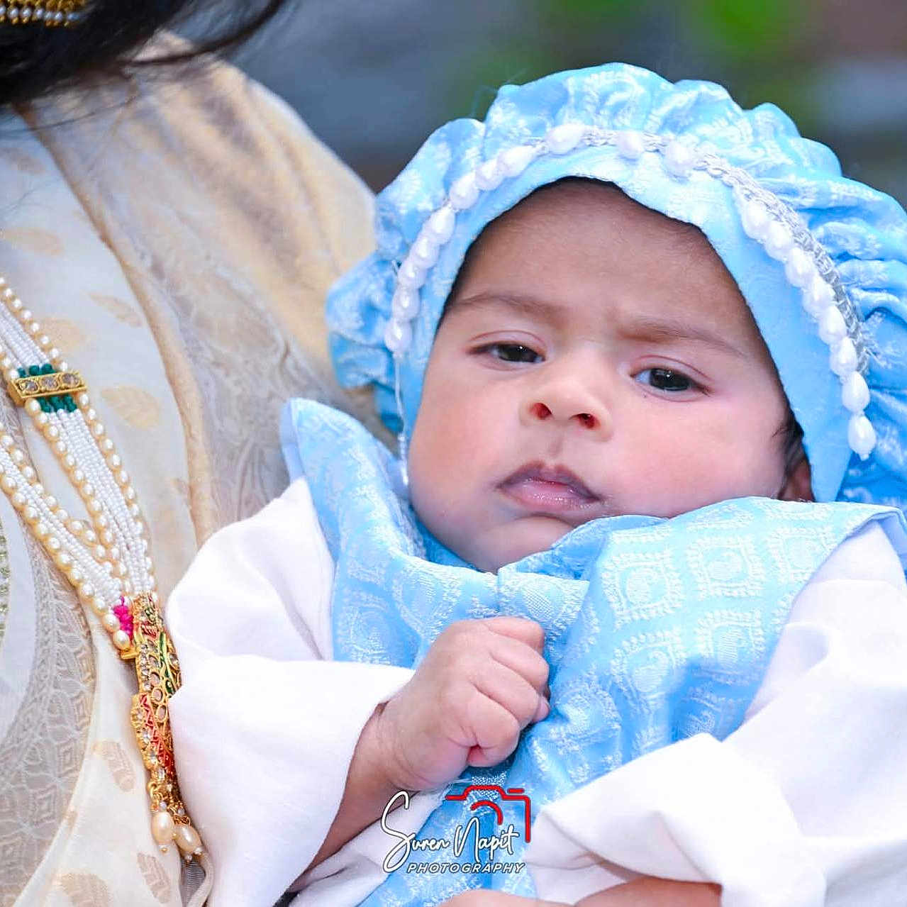 Samrat is registered to the contest to win money with this photo: baby, infant, child, portrait, blue_bonnet, white_clothing, adult, parent, holding, arms, face, eyes, indoor, traditional_clothing, jewelry, necklace, headwear, closeup, photography, family