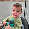toddler, child, sippy_cup, high_chair, indoor, kitchen, jars, gloves, green_sweater, curly_hair, cute, rosy_cheeks, person, seated, holding, food, snack, expression, face, young_child