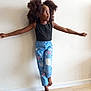 balance, barefoot, black_top, blue_jeans, butterfly_patches, casual_clothing, child, curly_hair, cute, expression, fashion, girl, indoor, playful, portrait, posing, standing, wall, wooden_floor, young