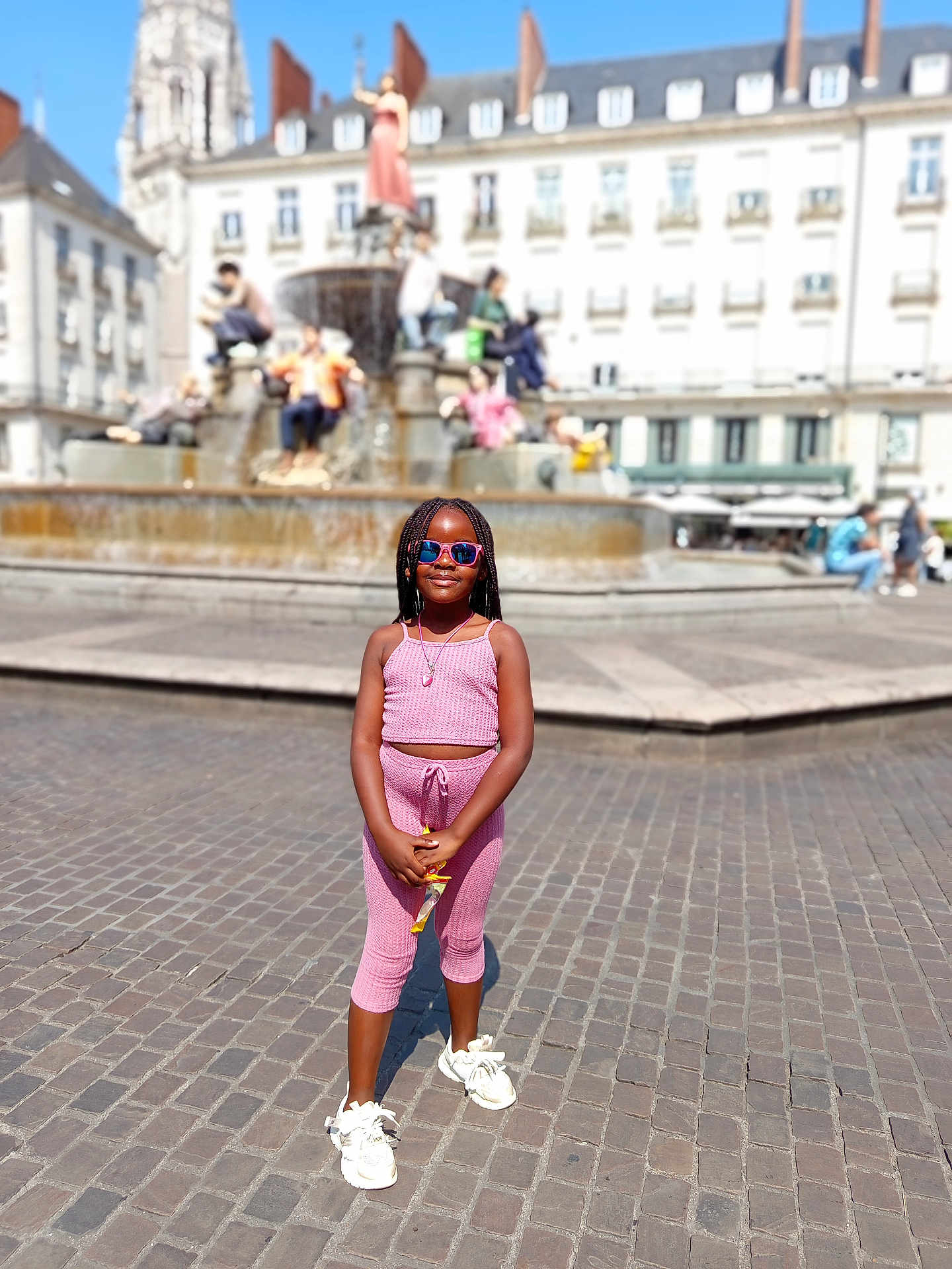 Tiana a rejoint le concours — aidez-le/la à gagner de superbes lots ! child, girl, pink_outfit, sunglasses, standing, outdoor, square, cobblestone, fountain, statue, buildings, sunny, daylight, casual_clothing, smiling, person, urban, youth, fashion, accessory