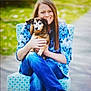 woman, dog, chair, blue_shirt, floral_pattern, jeans, outdoor, smiling, pet, animal, person, crossed_legs, holding, happy, casual, seat, greenery, blurred_background, companion, friendship