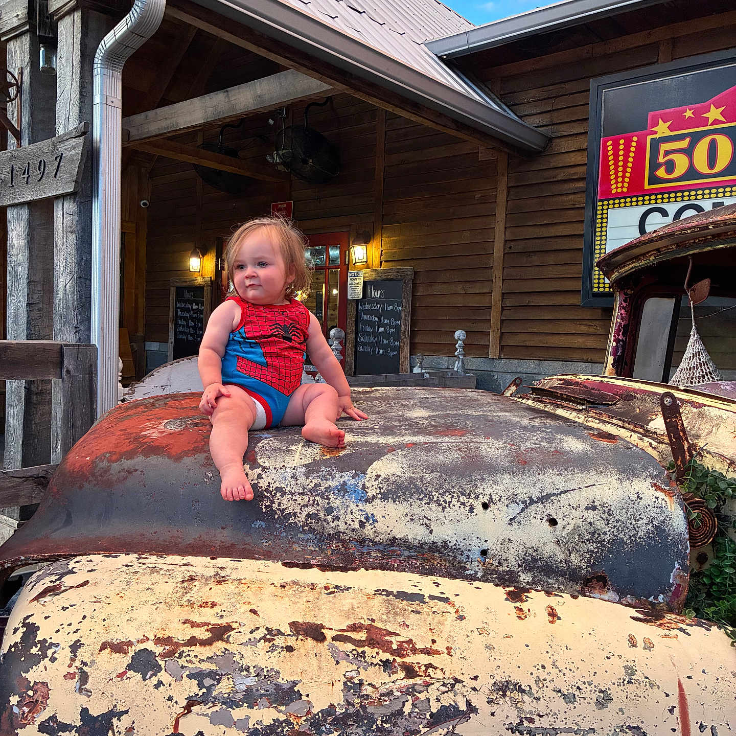 Caysen is registered to the contest to win money with this photo: baby, child, toddler, superhero_costume, spiderman, sitting, vintage_truck, truck_hood, rusty_vehicle, weathered_paint, rustic_building, wooden_building, outdoor, sunny, blue_sky, portrait, facial_expression, blonde_hair, automotive, nostalgic