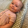baby, child, smiling, face, cushion, checkered_pattern, peach_dress, fruit_print, lying_down, soft_texture, indoors, cute, happy, infant, closeup, portrait, skin, head, eyes, gentle_lighting
