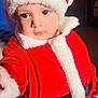 black_belt, child, christmas, curious_expression, cute, face, festive, holiday_costume, holiday_season, indoor, person, portrait, reaching_hand, red_clothing, santa_hat, soft_light, toddler, warm_colors, white_fur_trim, young_child