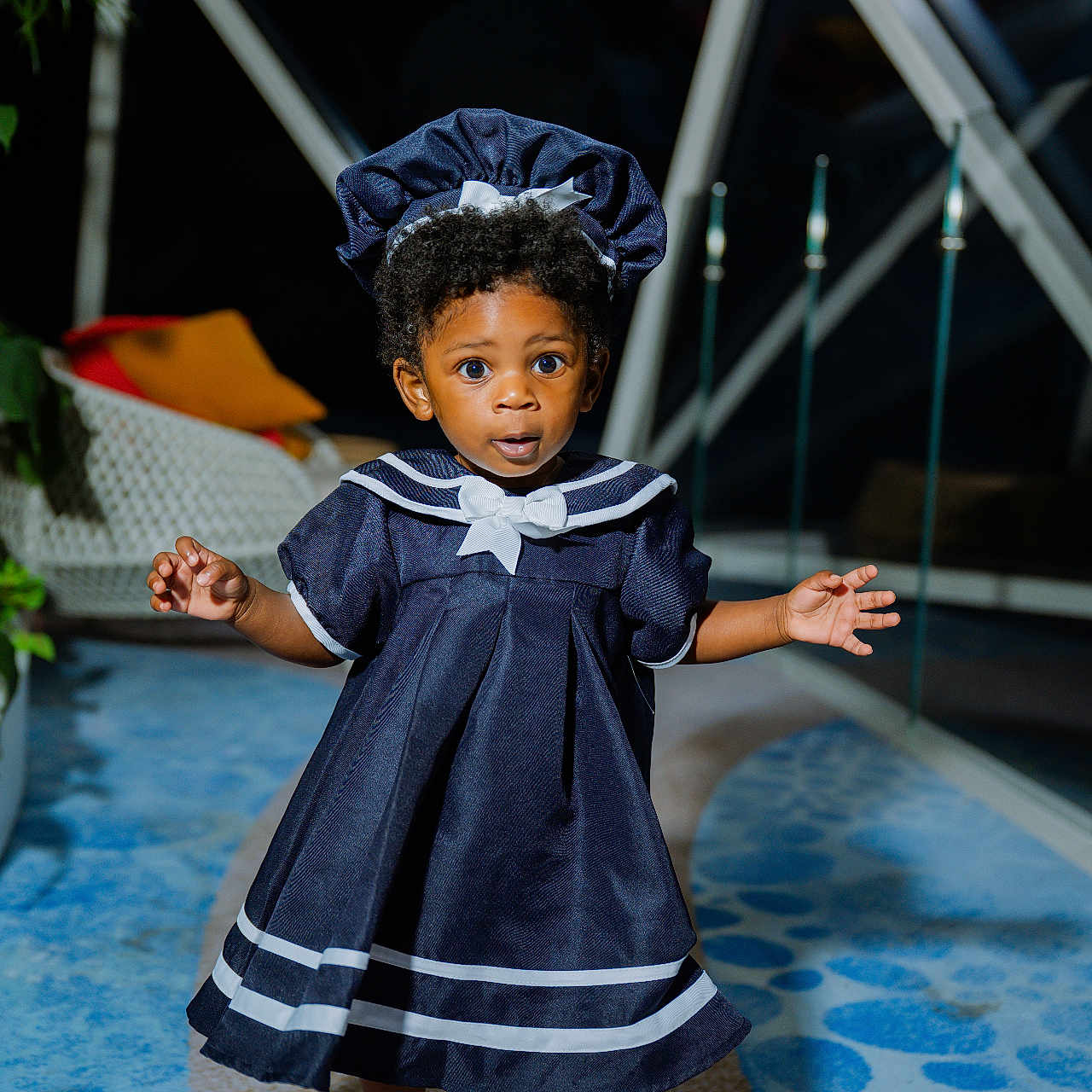 J'Alani is registered to the contest to win money with this photo: child, toddler, portrait, standing, sailor_dress, navy_blue, bonnet, curly_hair, wide_eyes, surprised_expression, outdoor_light, indoor, window, houseplant, carpet, sandals, fashion, full_body, smiling, adorable