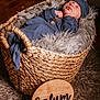 baby, newborn, sleeping, basket, woven_basket, blue_outfit, hat, fur_lining, shaggy_rug, wooden_plaque, name_sign, indoor, flooring, cozy, peaceful, infant, portrait, child, resting, cute