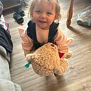 Bella is registered to the contest to win money with this photo: child, toddler, teddy_bear, toy, indoor, floor, wooden_floor, smiling, cute, person, messy_face, clothing, pink_sweater, overalls, home, living_room, footwear, slippers, table_leg, plastic_bag