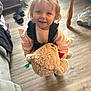 child, toddler, teddy_bear, toy, indoor, floor, wooden_floor, smiling, cute, person, messy_face, clothing, pink_sweater, overalls, home, living_room, footwear, slippers, table_leg, plastic_bag