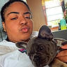 Coco is registered to the contest to win money with this photo: person, dog, french_bulldog, puppy, indoor, window, curtains, casual_clothing, white_shirt, earrings, hair_bun, hand, face, pet, couch, home, natural_light, closeup, relaxing, cozy