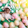 Kulture is registered to the contest to win money with this photo: baby, balloons, celebration, child, cute, festive, frilly, green_background, happy, hat, indoor, infant, pastel_colors, person, pink_dress, playful, portrait, sitting, smiling, soft_focus