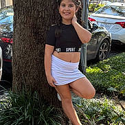 Camila is registered to the contest to win money with this photo: child, girl, smiling, braids, tree, outdoor, daylight, sporty_clothing, skirt, sneakers, greenery, plants, car, parking, nature, sunlight, happy, posing, casual, young