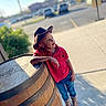 child, boy, cowboy_hat, red_shirt, jeans, boots, wooden_barrel, outdoor, daylight, sidewalk, parking_lot, greenery, casual_pose, leaning, portrait, sunlight, shadow, urban, thinking, young