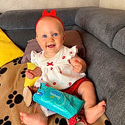 Lou a rejoint le concours — aidez-le/la à gagner de superbes lots ! baby, blanket, blue_eyes, child, couch, cute, footwear, happy, headband, indoors, infant, minnie_mouse_sandals, pampers_wipes, paw_prints, pillows, red_bow, red_shorts, sitting, smiling, white_dress